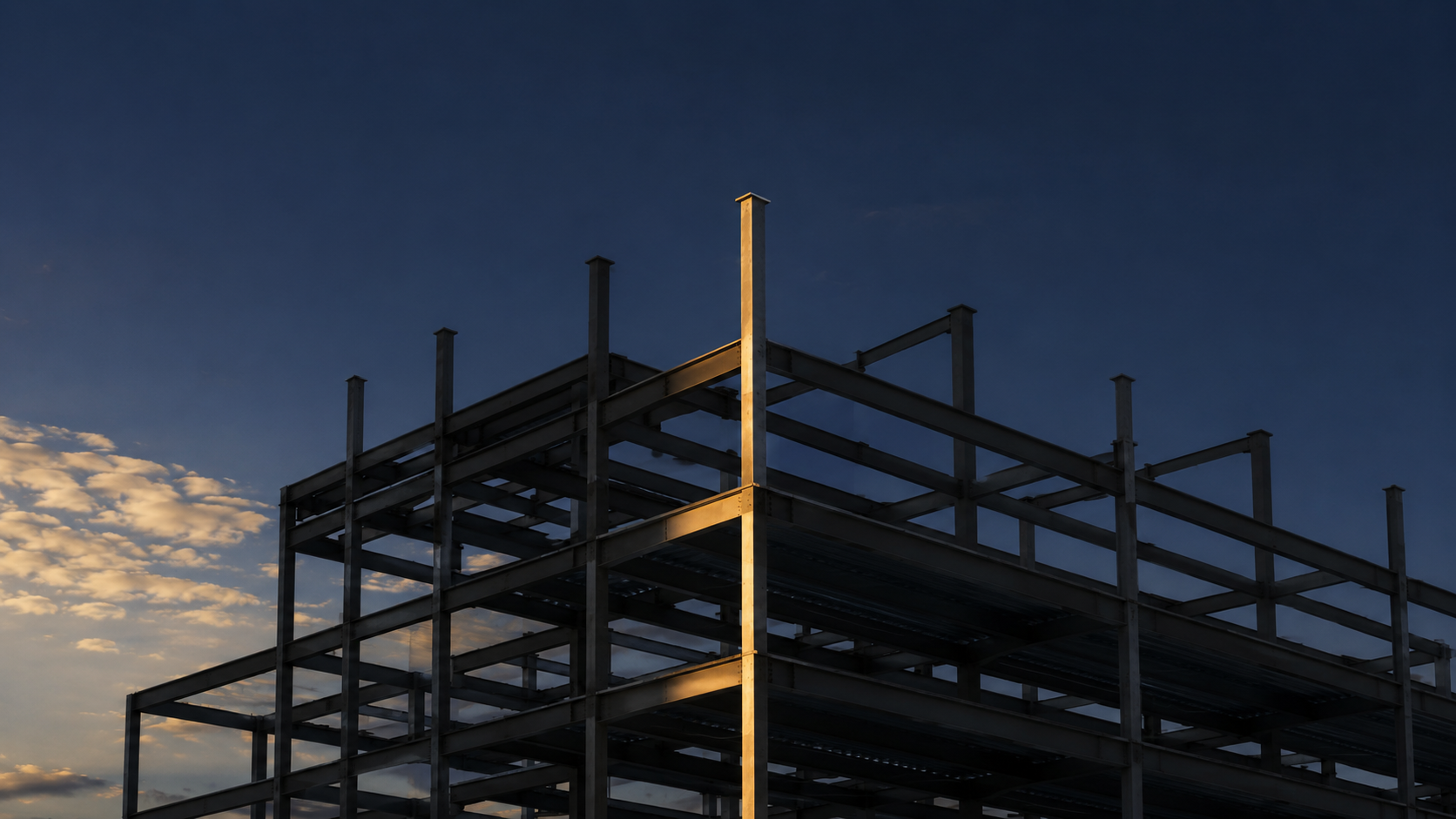 An unfinished steel building frame at dusk with a single vertical column catching the light, the rest of the structure in shadow.